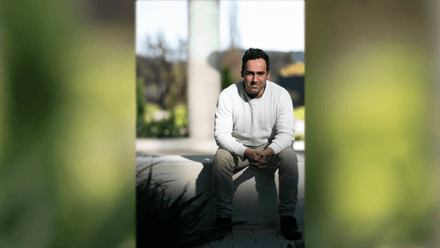 Portrait of Dr Filip Slaveski seated on an outdoor concrete bench. He is wearing a white cable-knit sweater and beige trousers, with his hands clasped and resting between his knees. He faces the camera with a neutral, attentive expression. The background is softly blurred with natural light and greenery visible, indicating a campus or garden setting.