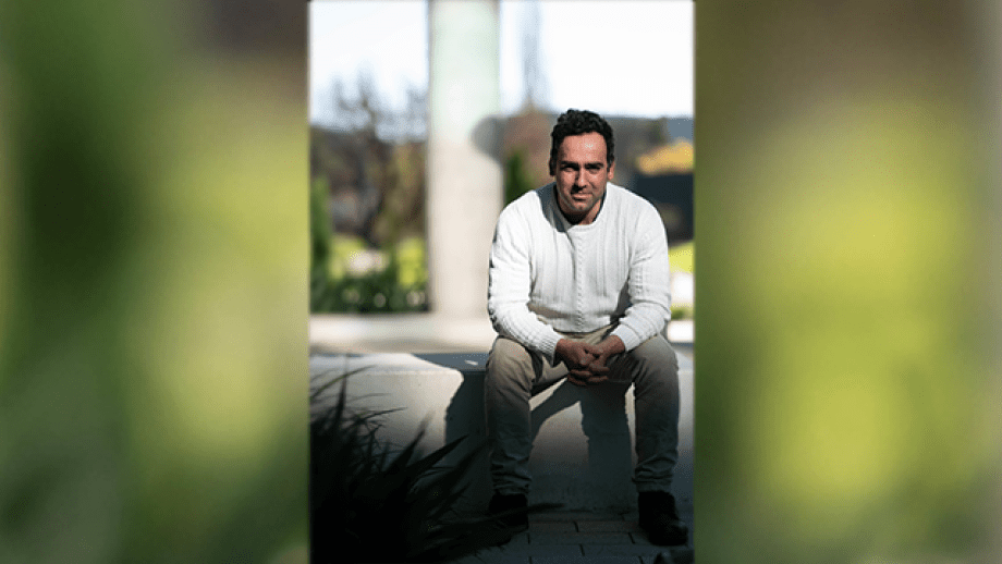 Portrait of Dr Filip Slaveski seated on an outdoor concrete bench. He is wearing a white cable-knit sweater and beige trousers, with his hands clasped and resting between his knees. He faces the camera with a neutral, attentive expression. The background is softly blurred with natural light and greenery visible, indicating a campus or garden setting.